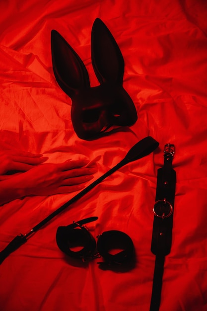 Conjunto de acessórios BDSM com mãos femininas está deitado na cama, máscara de algemas de chicote Flogger e gargantilha para sexo