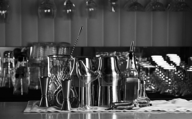 Conjunto de acessórios de bar para fazer coquetéis, localizado no bar, atrás está uma estante com licores e álcool forte. Mídia mista