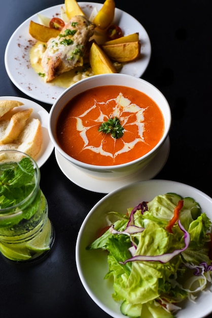 Conjunto de almoço feito de sopa de creme de tomate, salada, frango com batata, limonada em fundo preto