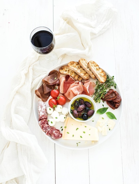 Conjunto de aperitivo de vinho Tomates cereja queijo parmesão variedade de carne fatias de pão tomates secos azeitonas e manjericão na placa de cerâmica redonda sobre o pano de fundo de madeira branca