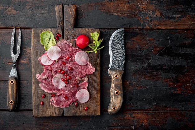 Conjunto de aperitivos frios de carpaccio de carne, com rabanete e granada, em uma tábua de servir de madeira, sobre uma velha mesa de madeira escura