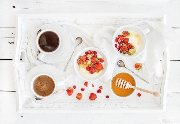 Conjunto de café da manhã saudável, mel de iogurte e tigelas de morango selvagem com café preto na bandeja de servir sobre o pano de fundo branco rústico de madeira