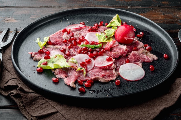 Conjunto de carpaccio de carne, com rabanete e granada, no prato, na velha mesa de madeira escura