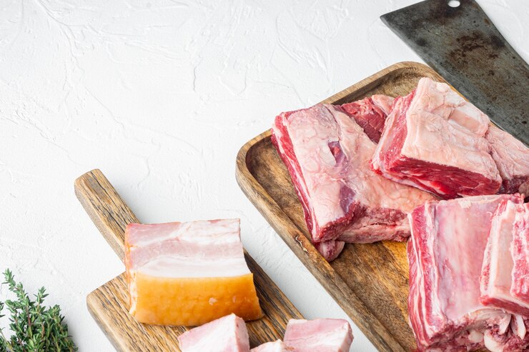 Foto conjunto de costelas curtas em guisado, com ingredientes, e velha faca de cutelo de açougueiro, sobre fundo de pedra branca, com espaço de cópia para o texto