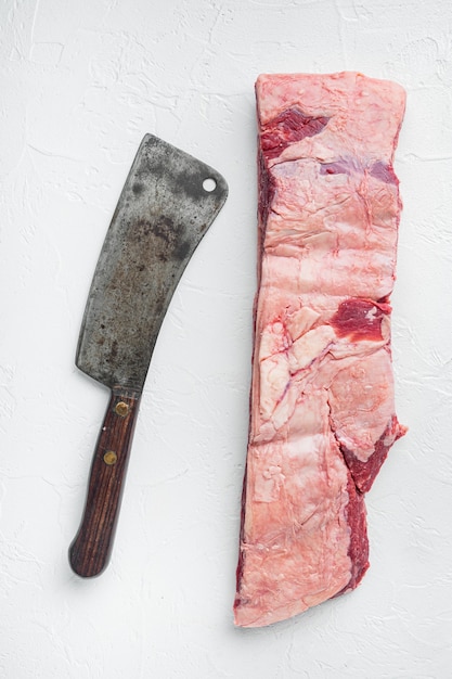 Conjunto de costelas de carne crua black angus em mármore e uma velha faca de cutelo de açougueiro, em fundo de pedra branca, vista de cima plana