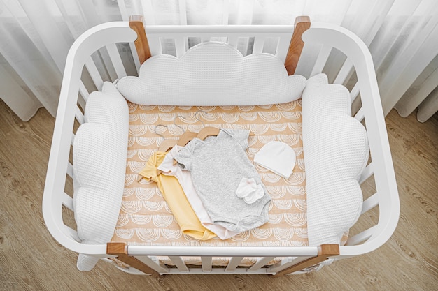 Foto conjunto de macacões de bebê para um recém-nascido no berço, berço. berço de madeira branco com almofadas em forma de nuvens no quarto do bebê. vista superior da cama da criança