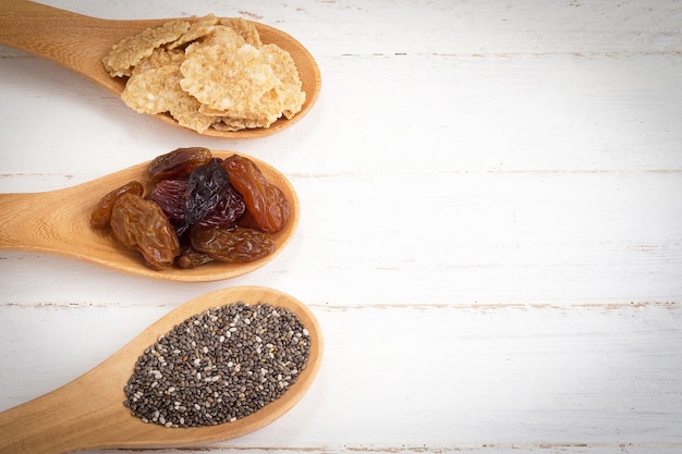 Conjunto de passas, flocos de cereais de trigo integral e sementes de chia em colher de madeira no fundo branco de madeira