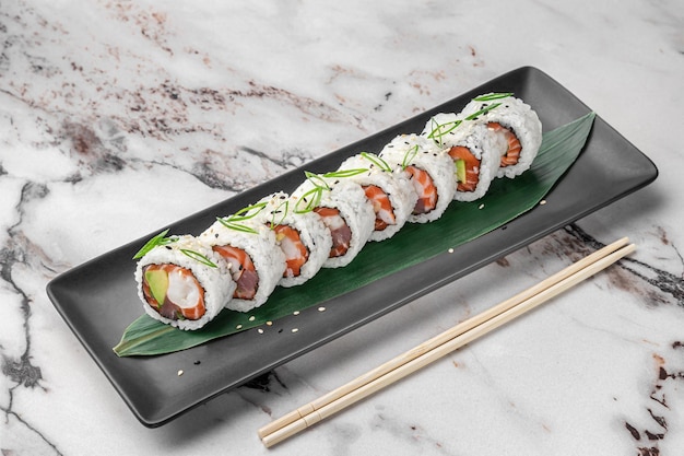 Conjunto de rolos de maki com arroz de abacate salmão atum camarão e folha de bambu verde em uma placa de cerâmica preta com pauzinho em uma vista lateral de fundo de mármore texturizado branco brilhante