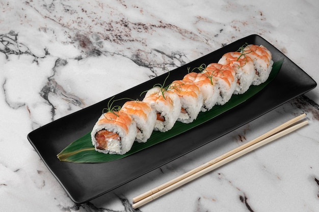 Conjunto de rolos de maki com microgreens de salmão de camarão tigre e folha de bambu verde em uma placa de cerâmica preta com pauzinho em uma vista lateral de fundo de mármore texturizado branco brilhante