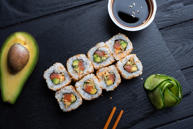 Conjunto de rolos de sushi com creme de queijo, arroz e salmão em uma placa preta decorada com molho de soja e abacate em uma mesa de madeira escura. Cozinha japonesa. Mesa de fotos de comida