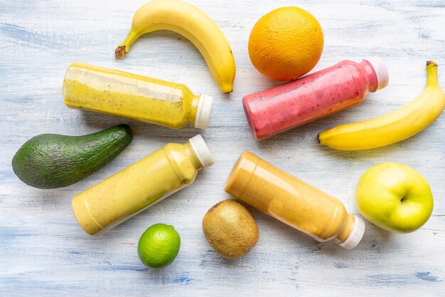 Conjunto de smoothies multi-coloridas em garrafas em composição com banana, kiwi, limão, abacate, sobre um fundo azul de madeira.