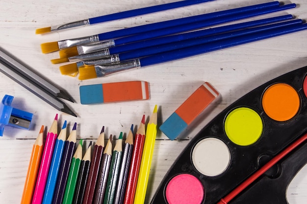 Conjunto de herramientas de dibujo sobre fondo blanco de madera. Pinturas de acuarelas, pinceles, lápices de colores, borradores y sacapuntas en el escritorio. Vista superior