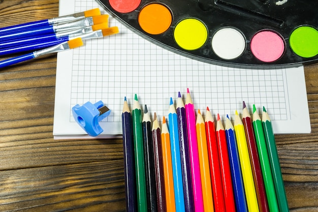 Conjunto de herramientas de dibujo sobre fondo de madera. Cuaderno en blanco, pinturas de acuarela, pinceles, lápices de colores en el escritorio