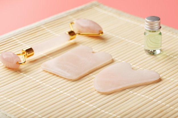 Un conjunto de herramientas para la técnica de masaje facial Gua Sha de cuarzo rosa natural. Rodillo, piedra de jade y aceite en un frasco de vidrio, sobre un fondo de paja para el cuidado de la cara y el cuerpo.
