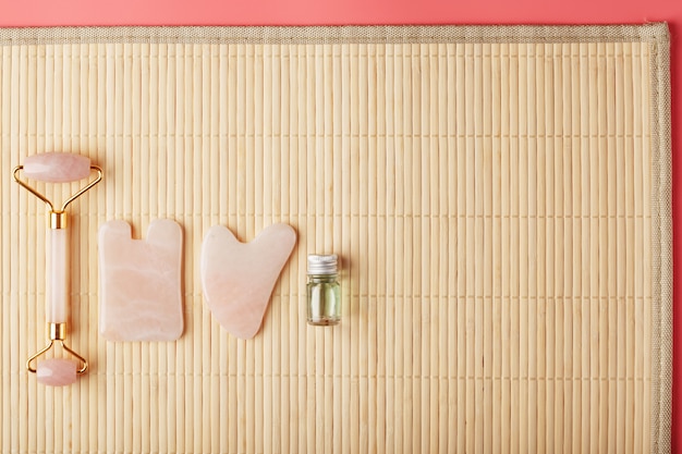 Un conjunto de herramientas para la técnica de masaje facial Gua Sha de cuarzo rosa natural. Rodillo, piedra de jade y aceite en un frasco de vidrio, sobre un fondo de paja para el cuidado de la cara y el cuerpo.