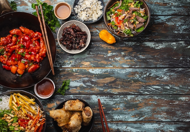 Conjunto de platos chinos variados en la mesa: pollo agridulce en sartén wok, dim sum, rollitos de primavera, fideos, ensalada, arroz, bollos al vapor. Cena o buffet de estilo asiático con vista superior y espacio para texto