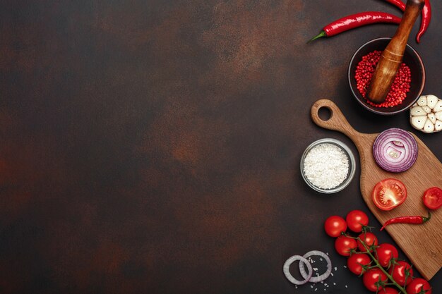 Conjunto de productos en la tabla de cortar de tomates cherry, romero, ajo, pimiento rojo, cebolla, sal y especias mortero sobre fondo marrón oxidado