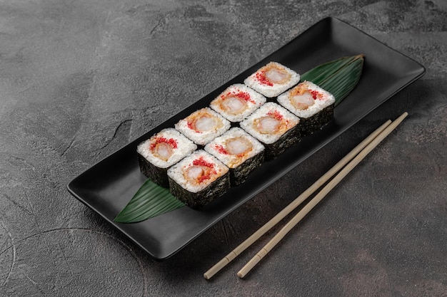 Conjunto de rollos de maki cuadrados con caviar de camarones de tigre y hojas de bambú verde en un plato de cerámica negra con palillos en una vista lateral de fondo con textura gris oscuro