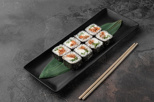 Conjunto de rollos de maki cuadrados con queso de cuajada de salmón frito y hoja de bambú verde en un plato de cerámica negra con palillos en una vista lateral de fondo con textura gris oscuro