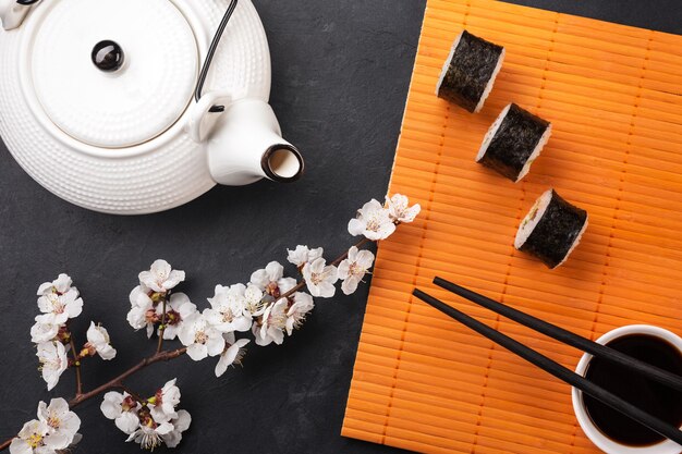 Conjunto de rollos de sushi y maki con rama de flores blancas y tetera en mesa de piedra. Vista superior.