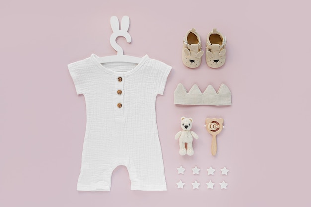Conjunto de ropa de bebé y complementos sobre fondo rosa. Body blanco en una linda percha con orejas de conejo y zapatos, corona de algodón y juguetes. Moda recién nacida. Endecha plana, vista superior