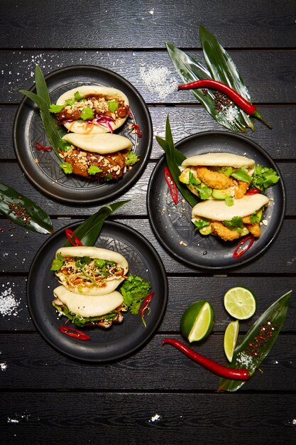 Un conjunto de tres platos asiáticos en platos negros sobre una mesa de madera decorada con limón, chile y harina. Apetitoso bao con verduras y carnes.