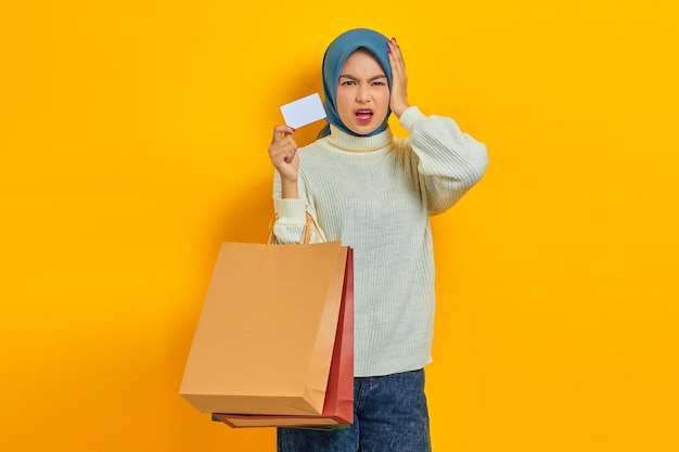 Conmocionada hermosa mujer asiática en suéter blanco sosteniendo bolsas de compras y tarjeta de crédito aislado sobre fondo amarillo