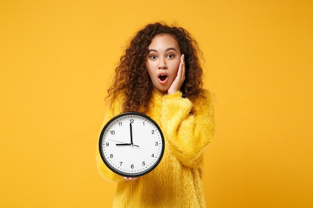 Conmocionada joven afroamericana en suéter de piel posando aislada sobre fondo amarillo naranja, retrato de estudio. Concepto de estilo de vida de las personas. Simulacros de espacio de copia. Sosteniendo el reloj, poniendo la mano en la mejilla.