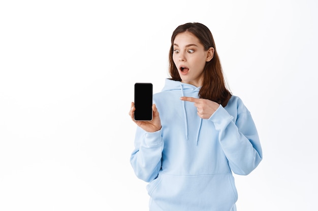 Conmocionada joven apuntando con el dedo a la pantalla de su teléfono, jadeando asombrada mientras mira la pantalla del teléfono inteligente, de pie contra la pared blanca