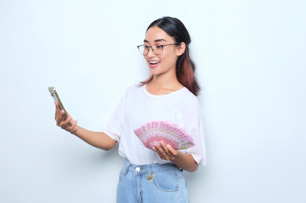 Conmocionada joven asiática con camiseta blanca usando teléfono móvil y sosteniendo billetes de dinero aislados en fondo blanco