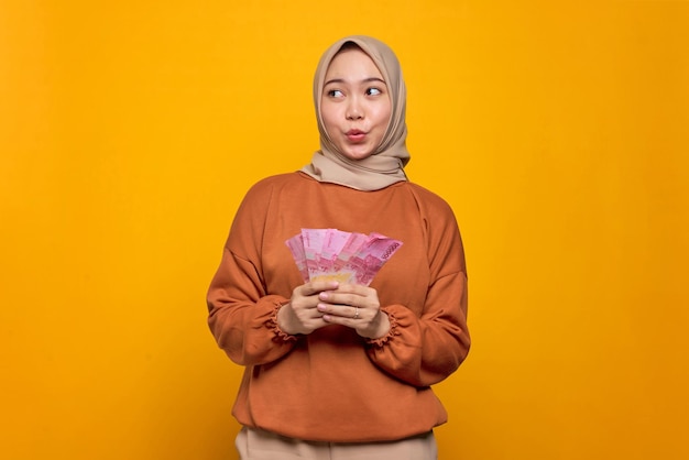 Conmocionada joven asiática con pantalones naranja sosteniendo billetes de dinero y mirando hacia arriba aislada sobre el fondo amarillo