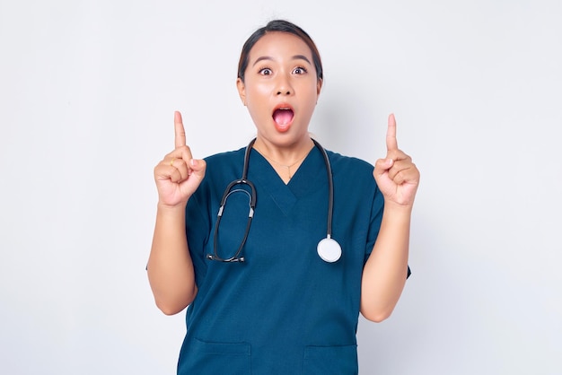 Conmocionada joven enfermera profesional asiática que trabaja con un uniforme azul apuntando con el dedo hacia el espacio de copia vacío aislado en fondo blanco Concepto de medicina sanitaria