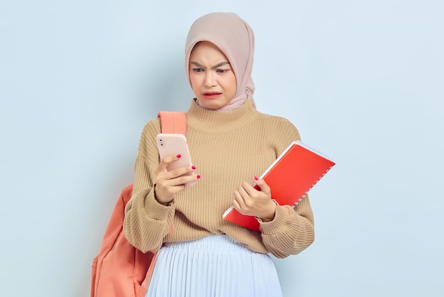 Conmocionada joven estudiante musulmana asiática en suéter marrón con mochila sosteniendo libro y teléfono móvil aislado sobre fondo blanco concepto de regreso a la escuela