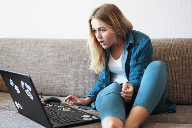 Conmocionada joven mirando la pantalla del portátil en casa viendo algo increíble en la computadora. Concepto de malas noticias. Pegatinas en una computadora portátil sin etiquetas