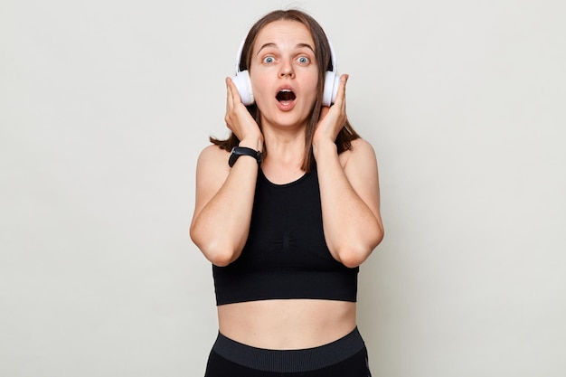 Conmocionada joven mujer caucásica en top negro escuchando música con auriculares mirando a la cámara aislada sobre fondo blanco mirando a la cámara con la boca abierta y ojos grandes