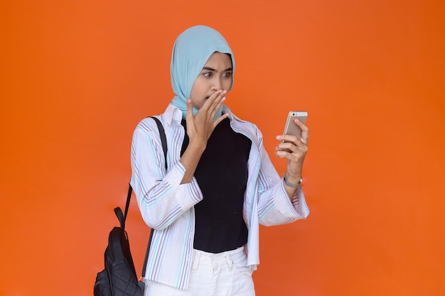 Conmocionada joven musulmana asiática mirando el teléfono inteligente mientras cubría la boca aislada sobre el bac naranja