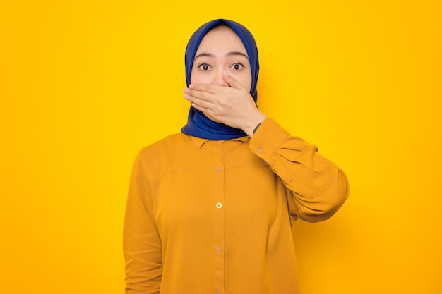 Conmocionada joven musulmana asiática vestida de naranja cubriendo la boca con la mano por error aislada de fondo amarillo