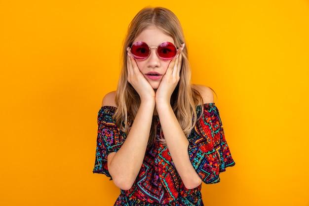 Conmocionada joven rubia eslava con gafas de sol poniendo las manos en la cara y mirando