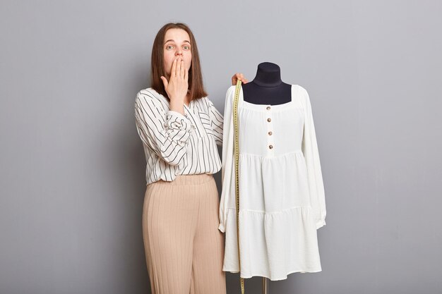 Conmocionada joven sastre adulta de pie cerca de maniquí con vestido nuevo aislado sobre fondo gris mirando a la cámara cubriendo la boca con la mano mostrando un nuevo atuendo increíble