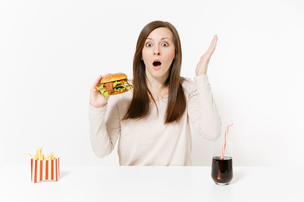 Conmocionada joven sentada a la mesa con hamburguesa, papas fritas, cola en botella de vidrio aislada sobre fondo blanco. Nutrición adecuada o comida rápida clásica estadounidense. Área de publicidad con espacio de copia.