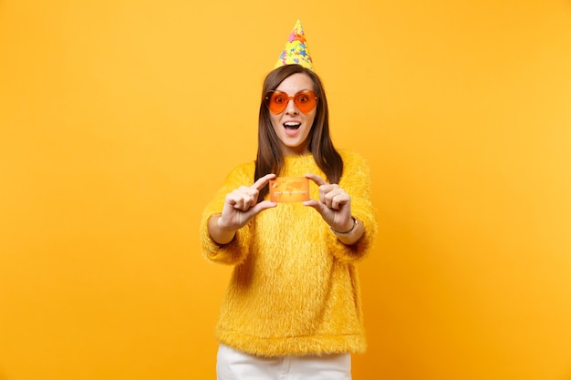 Conmocionada joven sorprendida en vasos de corazón naranja y sombrero de fiesta de cumpleaños con tarjeta de crédito, disfrutando de vacaciones, celebrando aislado sobre fondo amarillo. Personas sinceras emociones, concepto de estilo de vida.