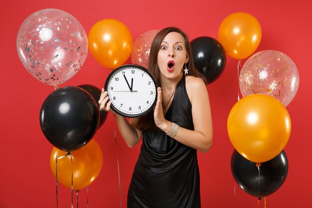 Conmocionada joven vestida de negro celebrando la celebración de reloj redondo en globo de aire de fondo rojo. Se acaba el tiempo. Día internacional de la mujer, feliz año nuevo, concepto de fiesta de vacaciones de maqueta de cumpleaños.