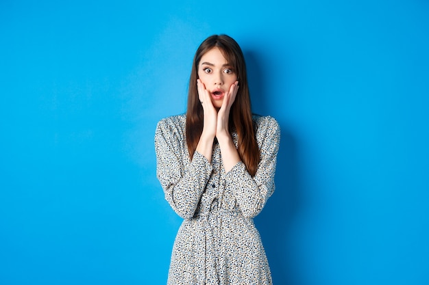 Conmocionada linda mujer jadeando sorprendida, caída de la mandíbula y mirando con incredulidad, de pie en vestido azul.