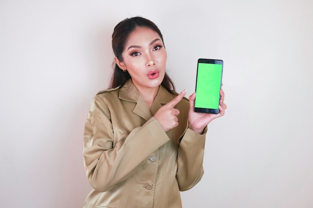 Conmocionada mujer asiática con uniforme marrón señalando con el dedo la pantalla verde en el teléfono inteligente en la mano