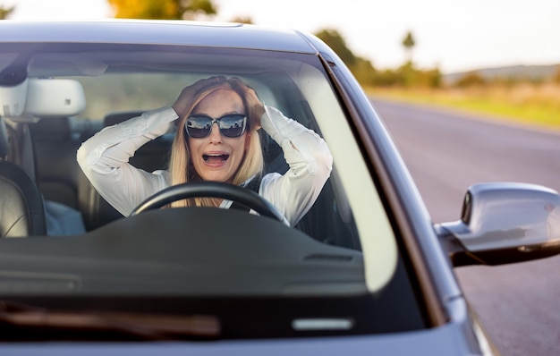 Conmocionada mujer caucásica con gafas de sol sentada en un auto con la boca abierta Conductora agarrándose la cabeza y gritando durante un accidente en la carretera