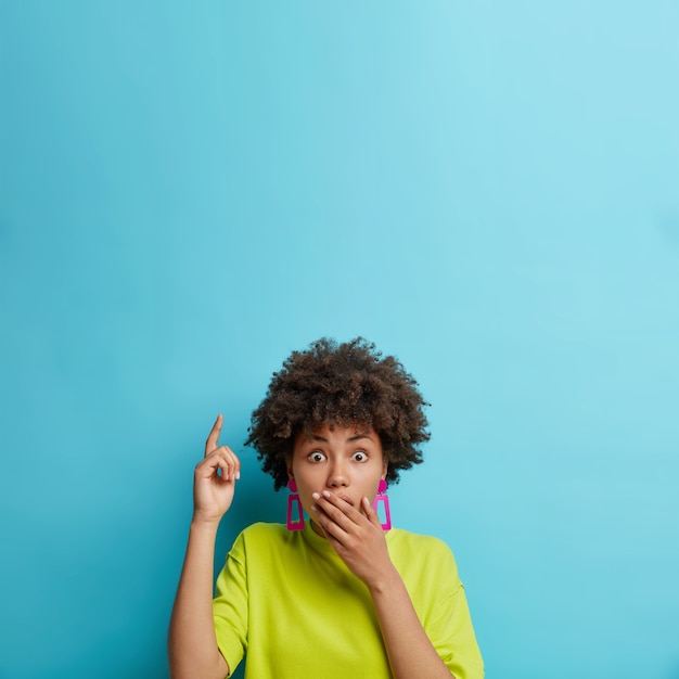 Conmocionada mujer de pelo rizado cubre los puntos de la boca con el dedo índice hacia arriba mira perpleja indica que la pancarta del logotipo plantea contra la pared azul