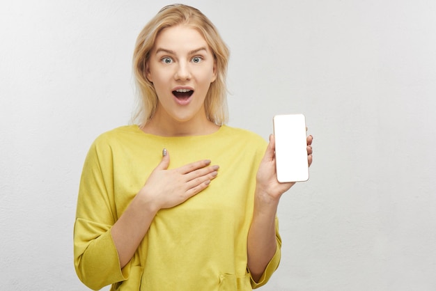 Conmocionada mujer rubia europea con teléfono móvil en la mano, ojos saltones de sorpresa, emociones de lo que ve en la pantalla, se queda boquiabierta. retrato, de, hermoso, niña, aislado, en, blanco, estudio