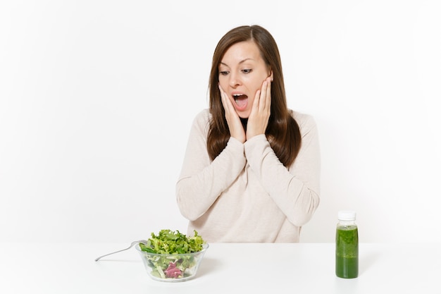 Conmocionada mujer sentada a la mesa con batidos de desintoxicación verde, ensalada en un recipiente de vidrio aislado sobre fondo blanco. Nutrición adecuada, comida vegetariana, estilo de vida saludable, concepto de dieta. Área con espacio de copia