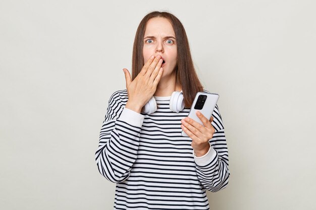 Conmocionada sorprendida mujer de cabello castaño con camisa a rayas posando aislada sobre fondo gris sosteniendo un teléfono inteligente cubriendo la boca con la palma asombrada por las noticias de Internet