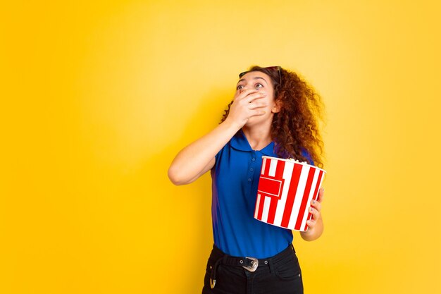 Foto conmocionado en el cine. retrato de niña de adolescentes caucásicos sobre fondo amarillo de estudio. modelo rizado femenino hermoso en camisa. concepto de emociones humanas, expresión facial, ventas, publicidad, educación. copyspace.